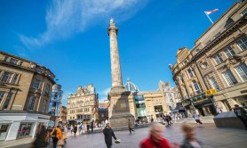 People walking down Newcastle’s Grey Street as Grey’s Monument tower overhead against a blue sky