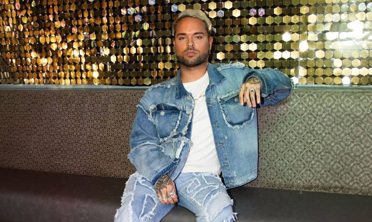 A man dressed in denim sitting on a sofa in front of a shiny gold wall